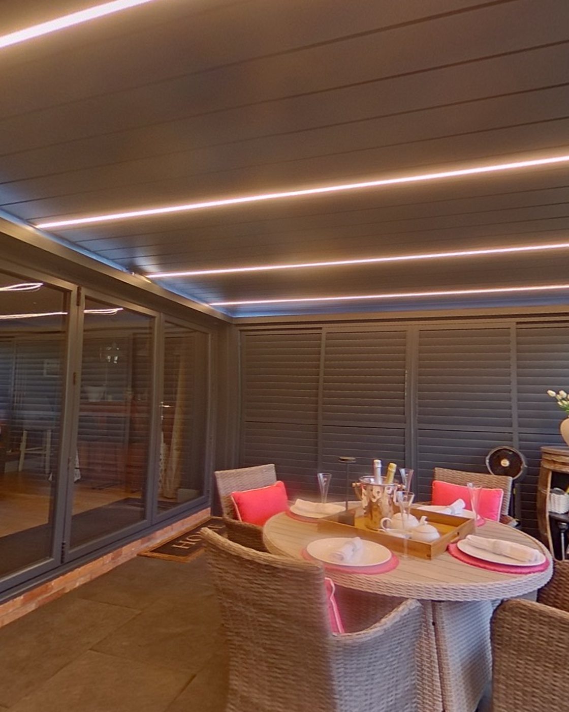 Our show pergola after dark, its bioclimatic louvres closed over a laid dining table and the integrated LED strips lit